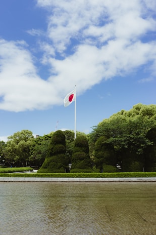 A flagpole with the Japanese flag waves against a backdrop of a clear blue sky and scattered white clouds. Below, manicured green bushes and trees are neatly arranged, with a paved path and a body of water in the foreground.