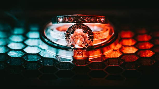 A dazzling diamond ring sits on top of a hexagonal patterned surface. The ring is elegantly crafted with a large, central diamond surrounded by smaller stones. The lighting creates a striking contrast with cool blue and warm red tones highlighting the metallic surface.