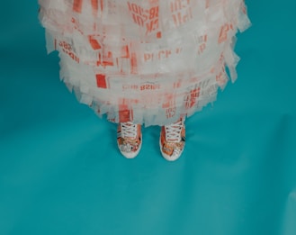 A person wearing a dress made from repurposed or recycled materials and colorful sneakers, standing on a solid teal-colored floor.