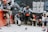 A group of workers take a break beside a wall covered with graffiti and posters. They wear construction attire, including helmets and boots, and some hold snacks or drinks. Two pigeons are on the ground in front of them.