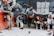 A group of workers take a break beside a wall covered with graffiti and posters. They wear construction attire, including helmets and boots, and some hold snacks or drinks. Two pigeons are on the ground in front of them.