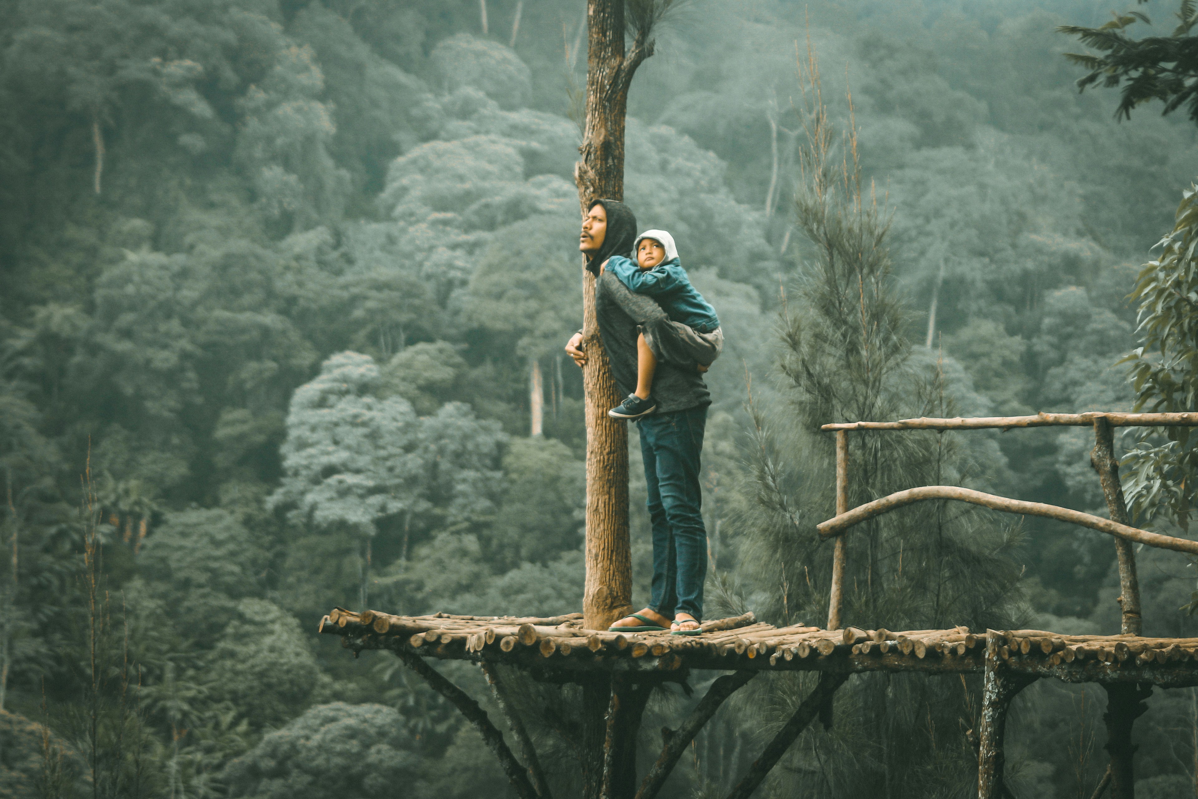 A man with a child on his back standing on a ledge looking at the view of a forest