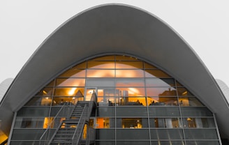 An arched modern building with a large glass facade, illuminated from inside with warm lights, exhibiting a futuristic architectural style. A staircase leads up to entrance doors framed by a series of rectangular window panes.