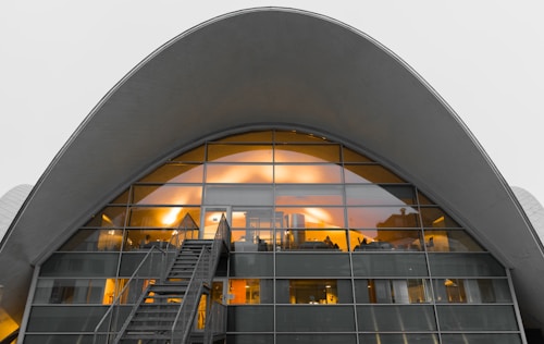 An arched modern building with a large glass facade, illuminated from inside with warm lights, exhibiting a futuristic architectural style. A staircase leads up to entrance doors framed by a series of rectangular window panes.