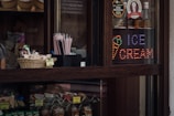 A dark wooden counter displays a variety of products including a basket of snacks and a set of pink straws. Bottles are lined up behind a glass partition, along with a colorful LED sign that reads 'Ice Cream' accompanied by an ice cream cone graphic.
