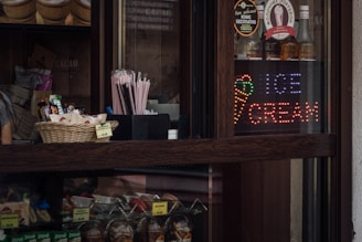 A dark wooden counter displays a variety of products including a basket of snacks and a set of pink straws. Bottles are lined up behind a glass partition, along with a colorful LED sign that reads 'Ice Cream' accompanied by an ice cream cone graphic.