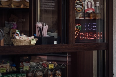 A dark wooden counter displays a variety of products including a basket of snacks and a set of pink straws. Bottles are lined up behind a glass partition, along with a colorful LED sign that reads 'Ice Cream' accompanied by an ice cream cone graphic.