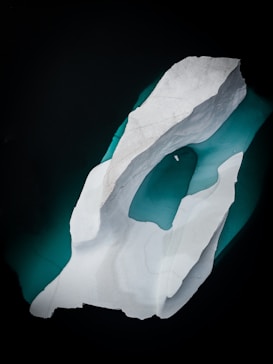 A large, jagged iceberg floats in dark water, casting a subtle reflection. The iceberg appears white with textured surfaces, and the surrounding water exhibits a deep teal color, creating a striking contrast.