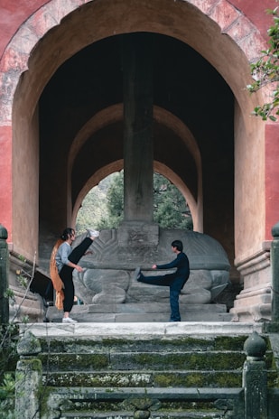 Two individuals are engaged in a martial arts practice beneath a large stone sculpture, standing on a set of moss-covered steps within an ancient arched structure. The background features a lush green environment visible through the archway.
