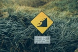 A yellow warning sign depicting a landslide hazard is placed amidst dense, lush green vegetation. Below the warning symbol, a smaller sign displays instructions in multiple languages, emphasizing a hazardous area that requires careful passage.