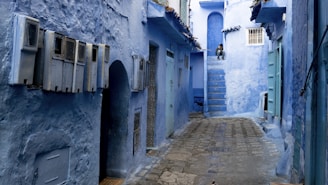 A narrow alleyway with walls and structures painted in varying shades of blue, characteristic of the city of Chefchaouen, Morocco. The cobblestone path leads into the distance, flanked by doors and windows. A person is sitting on steps at the end of the alley.