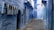 A narrow alleyway with walls and structures painted in varying shades of blue, characteristic of the city of Chefchaouen, Morocco. The cobblestone path leads into the distance, flanked by doors and windows. A person is sitting on steps at the end of the alley.