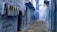 A narrow alleyway with walls and structures painted in varying shades of blue, characteristic of the city of Chefchaouen, Morocco. The cobblestone path leads into the distance, flanked by doors and windows. A person is sitting on steps at the end of the alley.