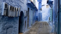 A narrow alleyway with walls and structures painted in varying shades of blue, characteristic of the city of Chefchaouen, Morocco. The cobblestone path leads into the distance, flanked by doors and windows. A person is sitting on steps at the end of the alley.