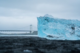 A large, clear blue ice block lies on a dark, textured surface with a bridge in the background. The sky appears overcast, creating a moody contrast between the icy blue of the block and the gray surroundings.