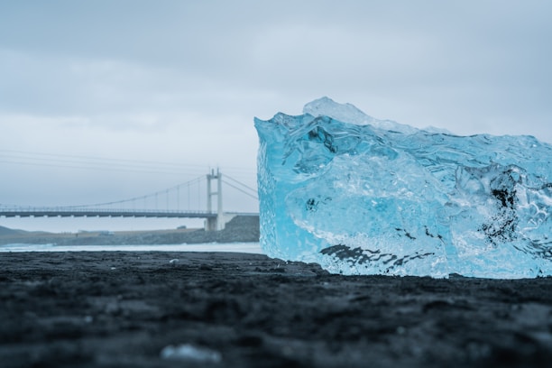 A large, clear blue ice block lies on a dark, textured surface with a bridge in the background. The sky appears overcast, creating a moody contrast between the icy blue of the block and the gray surroundings.