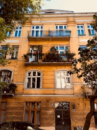 A four-story residential building with a warm yellow facade features multiple windows and balconies adorned with a variety of potted plants and flowers. The architecture includes arched and rectangular windows, and the building is surrounded by leafy green trees. A black wrought iron gate serves as the main entrance at street level.