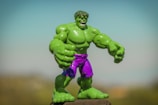 A green superhero action figure with large muscles and a fierce expression stands prominently. It is wearing purple shorts and is positioned against a blurred, natural background with a clear blue sky above.
