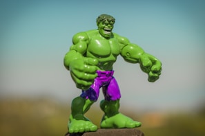 A green superhero action figure with large muscles and a fierce expression stands prominently. It is wearing purple shorts and is positioned against a blurred, natural background with a clear blue sky above.