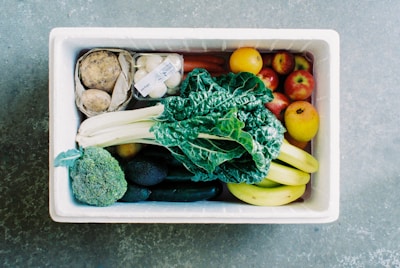A white box filled with a variety of fresh produce, including leafy greens, bananas, apples, cucumbers, broccoli, eggs in a carton, potatoes, and other fruits and vegetables. The contents are neatly arranged and display a diverse selection of colors and textures.