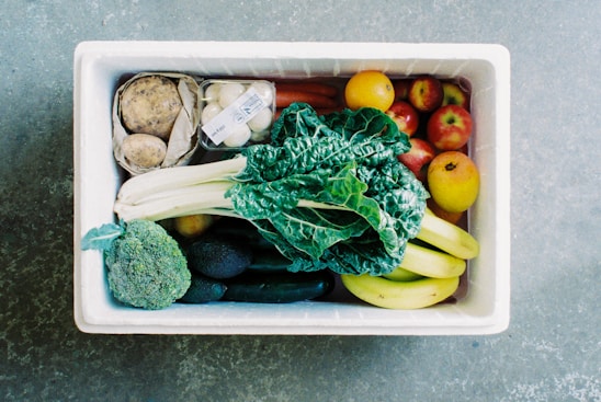 A white box filled with a variety of fresh produce, including leafy greens, bananas, apples, cucumbers, broccoli, eggs in a carton, potatoes, and other fruits and vegetables. The contents are neatly arranged and display a diverse selection of colors and textures.