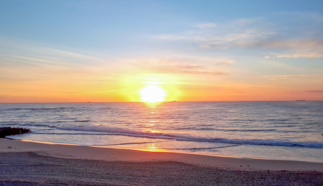 A serene beach scene at sunrise with the sun rising over the horizon, casting warm hues across the sky and reflecting off the calm ocean waves. The sandy beach stretches across the foreground with a clear gradient of light and shadow, while gentle waves lap against the shore.