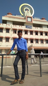 A young man in a blue shirt stands casually against a railing in front of a large building. The building has a prominent clock and signs in multiple languages, indicating it is a railway station in Varanasi.