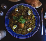 A vibrant blue ceramic plate holds a richly spiced dish featuring cooked chicken, whole green lentils, and small halved boiled eggs. A sprig of fresh mint sits atop the dish, enhancing its presentation. The plate is accompanied by utensils and partially visible slices of bread, with salt and pepper shakers nearby.