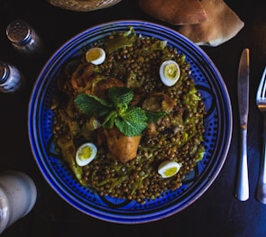 A vibrant blue ceramic plate holds a richly spiced dish featuring cooked chicken, whole green lentils, and small halved boiled eggs. A sprig of fresh mint sits atop the dish, enhancing its presentation. The plate is accompanied by utensils and partially visible slices of bread, with salt and pepper shakers nearby.