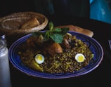 A blue plate is filled with a dish consisting of legumes, pieces of chicken, and garnished with mint leaves. Two pieces of boiled egg are placed on top of the dish. In the background, a wicker basket contains pieces of bread. A glass container, possibly for condiments or sauce, is seen on the side.