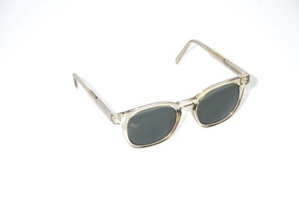 A pair of sunglasses with transparent frames and dark lenses is positioned at an angle against a light background. The frames have a subtle golden hue and the design is classic, featuring a rectangular shape.