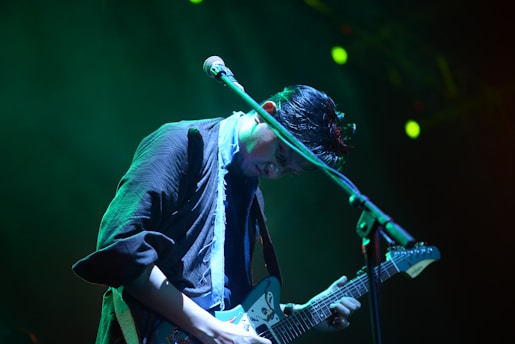A musician plays an electric guitar under stage lighting, creating an atmospheric and immersive setting. The dim lighting casts cool blue and green tones across the scene, highlighting the musician's focus and concentration.