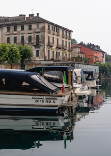 Several boats are docked in a calm body of water, reflecting the scene around them. An old, weathered stone building with a rustic charm stands by the waterfront, and a restaurant sign is visible. The scene suggests a quaint, historic setting, possibly in a small town. Trees and more colorful buildings are visible in the background, enhancing the picturesque quality.