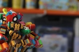 A colorful collection of decorative pencil toppers featuring various shapes such as monkeys, a car, a smiling face, and a heart. These toppers sit atop pencils in a vibrant array of colors against a blurred background.