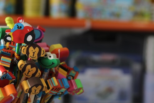 A colorful collection of decorative pencil toppers featuring various shapes such as monkeys, a car, a smiling face, and a heart. These toppers sit atop pencils in a vibrant array of colors against a blurred background.