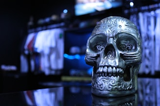 A metallic skull with intricate engravings is prominently displayed against a blurred background of clothing racks. The skull features detailed carvings and has a shiny, reflective surface. The setting is dimly lit, with a bluish tint highlighting the skull and the surrounding area.