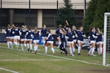 A group of women in navy blue and white athletic uniforms are jogging together on a soccer field. They are wearing matching tops with a specific logo and white shorts. The background includes a fence, some trees, and a goal post on the right side.