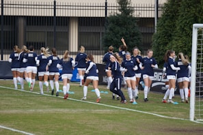 A group of women in navy blue and white athletic uniforms are jogging together on a soccer field. They are wearing matching tops with a specific logo and white shorts. The background includes a fence, some trees, and a goal post on the right side.