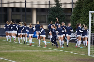 A group of women in navy blue and white athletic uniforms are jogging together on a soccer field. They are wearing matching tops with a specific logo and white shorts. The background includes a fence, some trees, and a goal post on the right side.