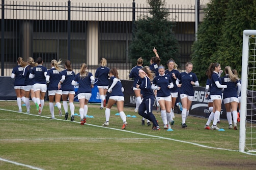 A group of women in navy blue and white athletic uniforms are jogging together on a soccer field. They are wearing matching tops with a specific logo and white shorts. The background includes a fence, some trees, and a goal post on the right side.