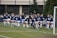 A group of women in navy blue and white athletic uniforms are jogging together on a soccer field. They are wearing matching tops with a specific logo and white shorts. The background includes a fence, some trees, and a goal post on the right side.