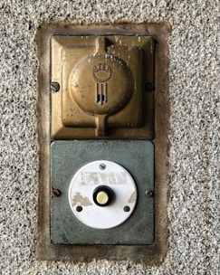 A vintage wall-mounted electrical outlet and a round doorbell button are affixed to a rough-textured concrete wall. The outlet, made of metallic and bronze tones, features a weathered appearance with an embossed logo. Below it, a circular white button, showing signs of wear, has a yellow center and is secured with screws.
