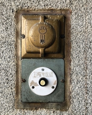 A vintage wall-mounted electrical outlet and a round doorbell button are affixed to a rough-textured concrete wall. The outlet, made of metallic and bronze tones, features a weathered appearance with an embossed logo. Below it, a circular white button, showing signs of wear, has a yellow center and is secured with screws.
