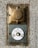 A vintage wall-mounted electrical outlet and a round doorbell button are affixed to a rough-textured concrete wall. The outlet, made of metallic and bronze tones, features a weathered appearance with an embossed logo. Below it, a circular white button, showing signs of wear, has a yellow center and is secured with screws.