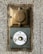 A vintage wall-mounted electrical outlet and a round doorbell button are affixed to a rough-textured concrete wall. The outlet, made of metallic and bronze tones, features a weathered appearance with an embossed logo. Below it, a circular white button, showing signs of wear, has a yellow center and is secured with screws.
