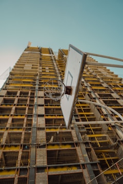 A construction site features a partially built high-rise building with exposed metal beams and concrete structures. In the foreground, a basketball hoop is prominently visible, highlighting a contrast between leisure and work environments.