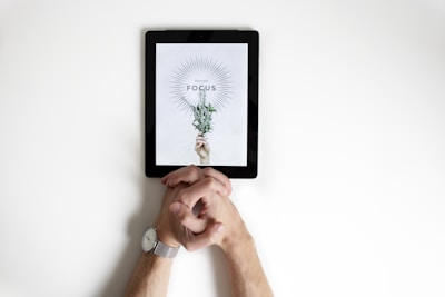A digital tablet placed on a white surface displays an image of a hand holding a small bouquet of flowers. Above the bouquet, the word 'FOCUS' is prominently featured with a radiating line design surrounding it. In the foreground, a person's hands are clasped together, resting on the surface, with a watch visible on one wrist.