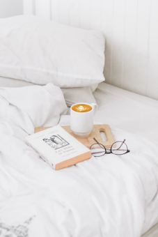 A cozy bedroom scene featuring a neatly made bed with crisp white linens. On the bed, there is a wooden tray holding a mug of latte art coffee, a paperback book, and a pair of eyeglasses. The ambiance is tranquil and inviting.