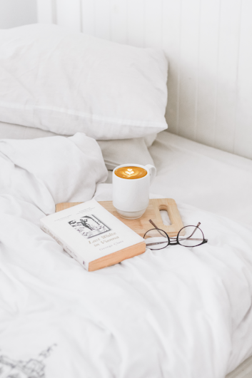 A cozy bedroom scene featuring a neatly made bed with crisp white linens. On the bed, there is a wooden tray holding a mug of latte art coffee, a paperback book, and a pair of eyeglasses. The ambiance is tranquil and inviting.