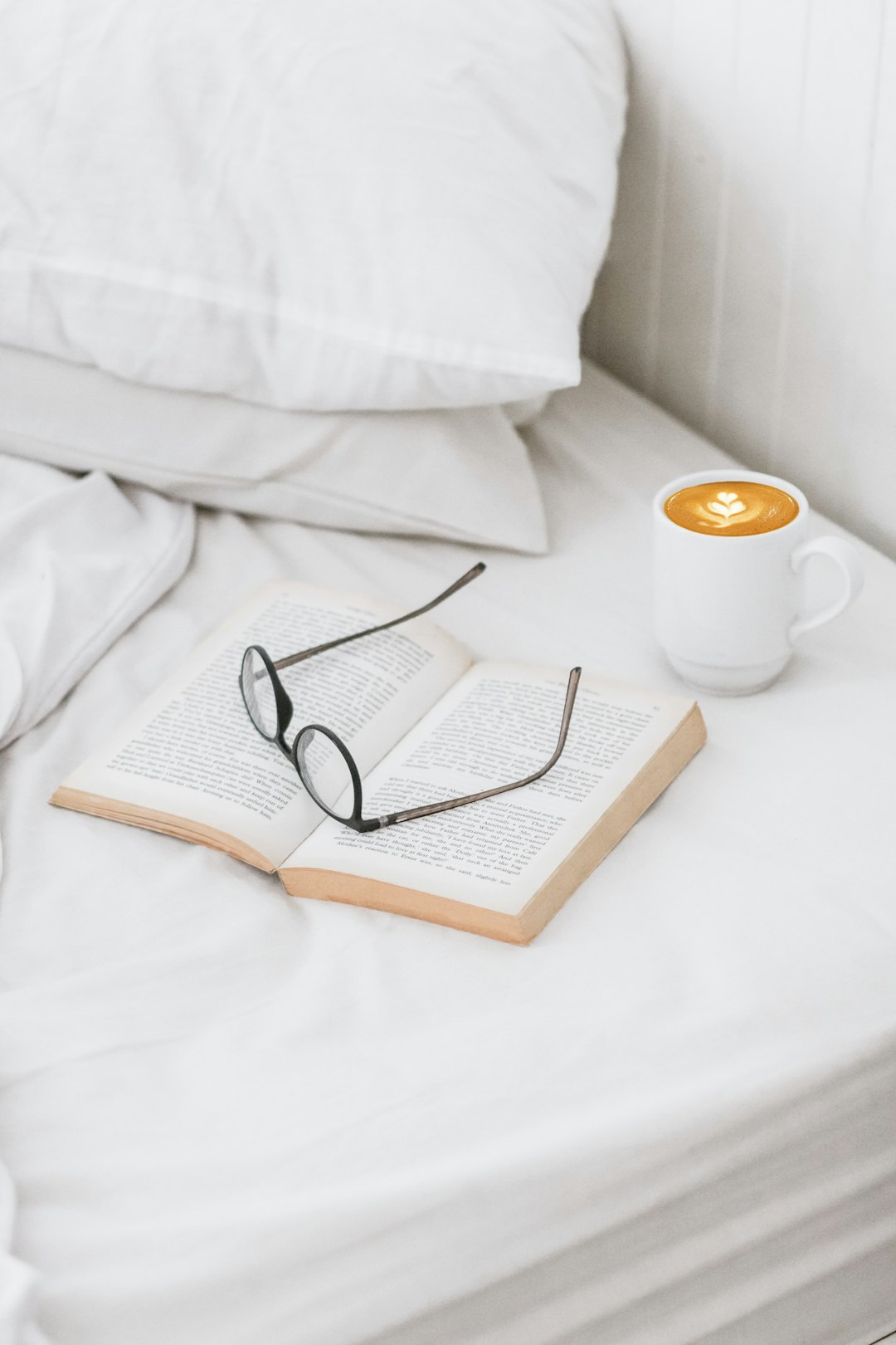  coffee on ceramic mug, book, and eyeglass on bed sheet